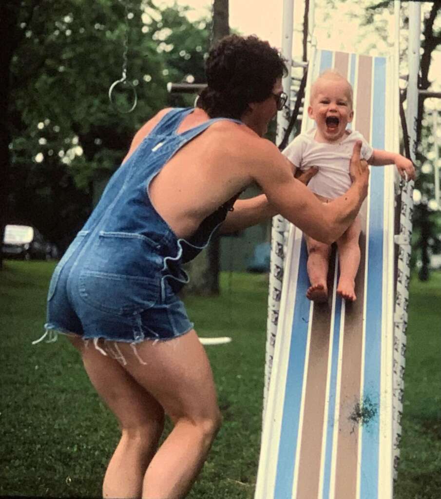 An adult in overalls is helping a laughing baby slide down a playground slide. They are outdoors on a grass field with trees in the background. The baby is wearing a white onesie and seems excited and happy, while the adult is smiling and supporting the baby.