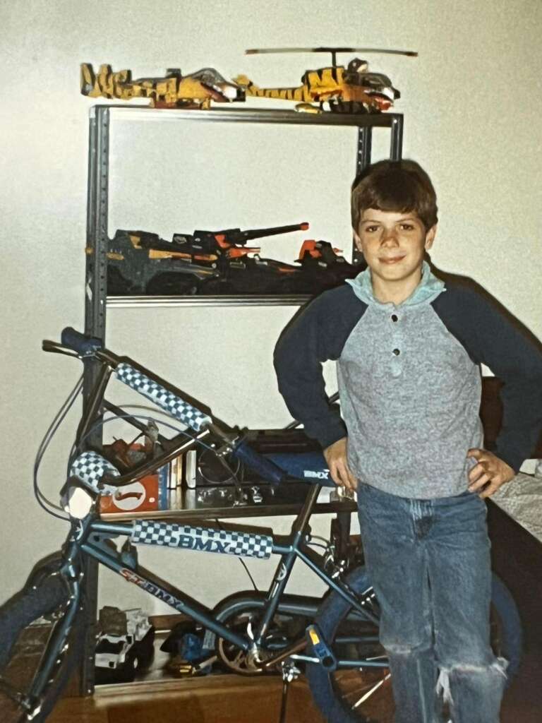 A young boy smiles while standing next to a blue BMX bike with checkered decals. He's wearing a gray and blue long-sleeve shirt and jeans. Behind him is a metal shelf with various toy models, including helicopters and planes.