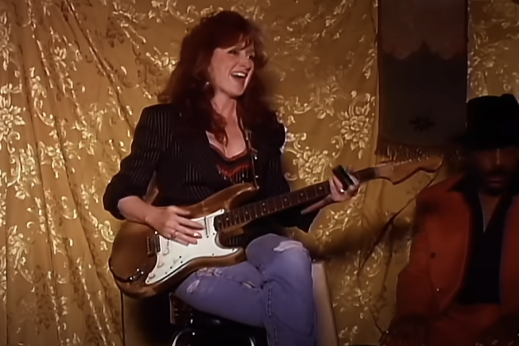 A woman with long, red hair plays an electric guitar while smiling. She is wearing a black pinstripe blazer and sits on a stool against a gold, patterned backdrop. Another person in an orange outfit and hat is partially visible nearby. The mood appears lively and relaxed.