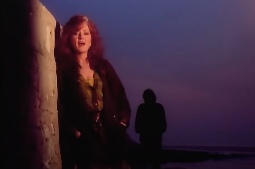 A woman with wavy red hair stands against a wall at dusk, singing passionately. She wears a dark jacket over a green shirt. In the background, a silhouette of a person walking on a beach is visible against the twilight sky.
