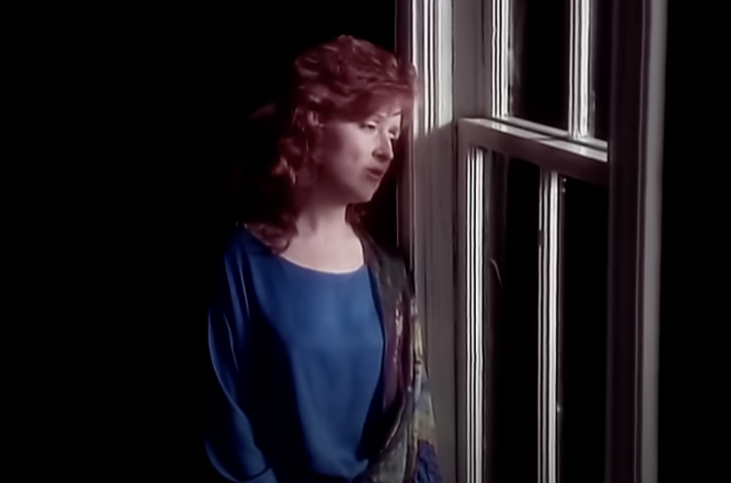 A person with curly red hair wearing a blue blouse is standing by a window, looking contemplative. The background is dark, and they are partially illuminated by the natural light coming through the window.