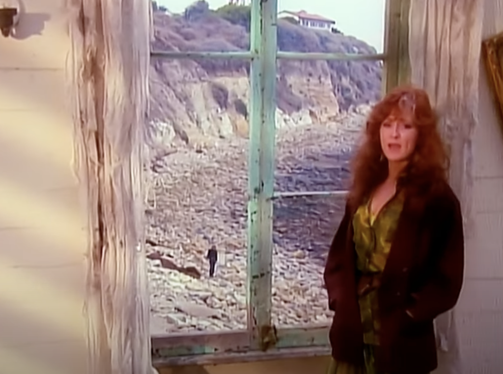 A woman with long, curly hair stands inside a rustic building near a weathered window with tattered curtains. Through the window, a rocky coastline and a house atop a cliff are visible, along with a lone figure walking along the shore.