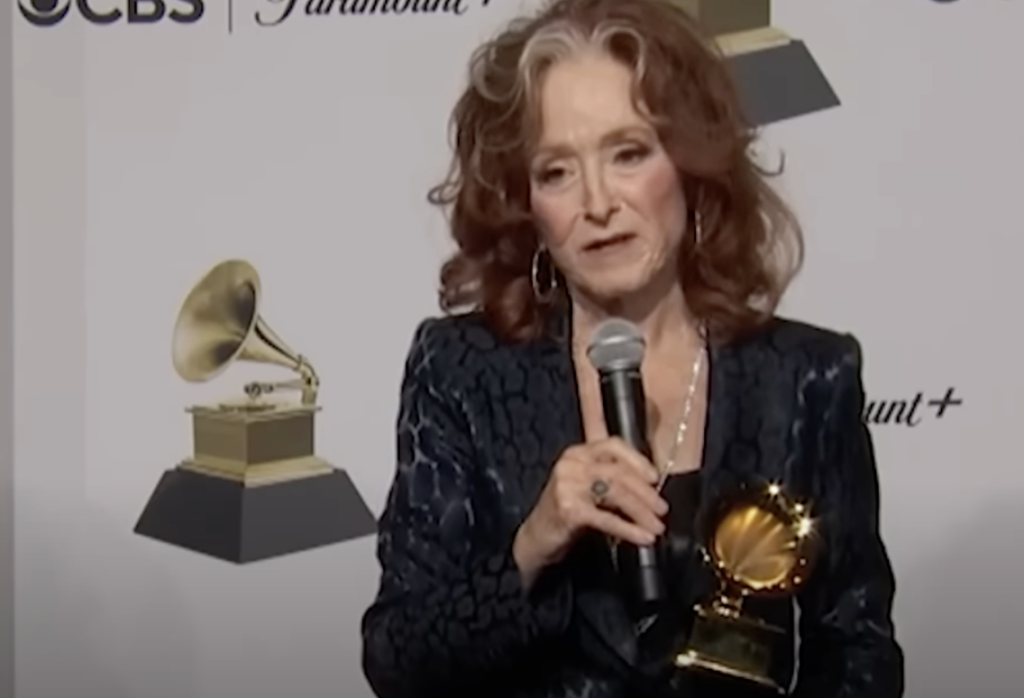 A person with red curly hair, wearing a dark patterned suit, is holding a Grammy award and speaking into a microphone. The background features the Grammy Awards logos and CBS and Paramount+ branding.