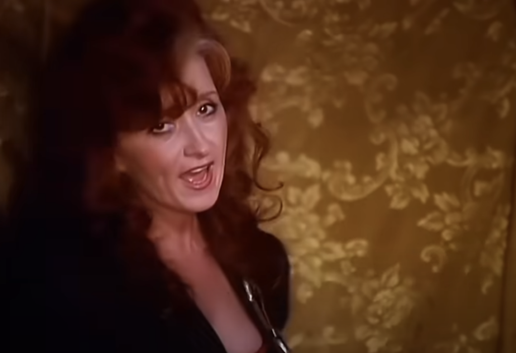 A woman with long, curly red hair and a black outfit sings against a yellow, floral-patterned backdrop. She appears to be performing with a lively expression.