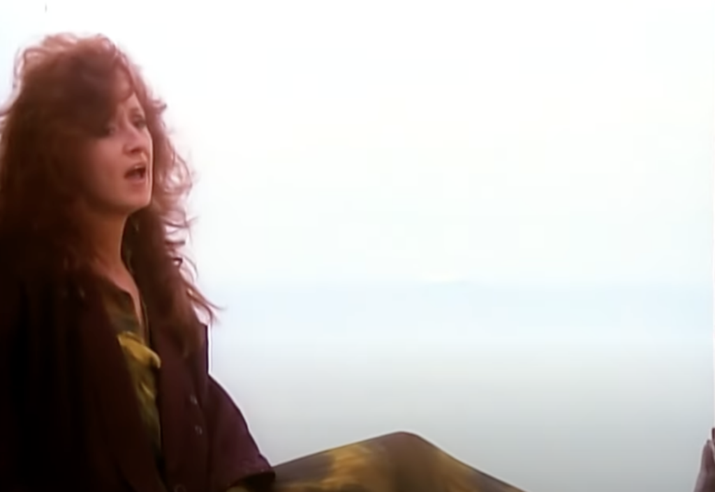 A woman with curly, reddish-brown hair sings passionately. She is wearing a dark-colored outfit and sits against the backdrop of a cloudy sky. The image conveys a sense of emotion and intensity.