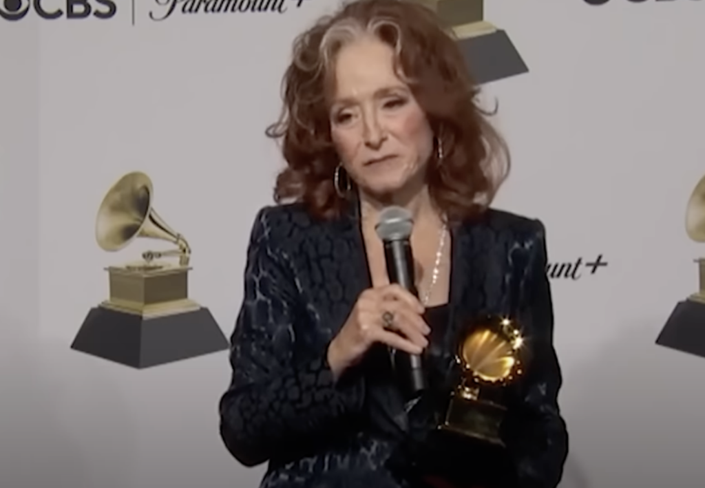 A woman with red hair, wearing a dark blue textured outfit, holds a Grammy award in one hand and speaks into a microphone in the other. She is standing in front of a backdrop featuring Grammy logos, CBS, Paramount+, and other branding.