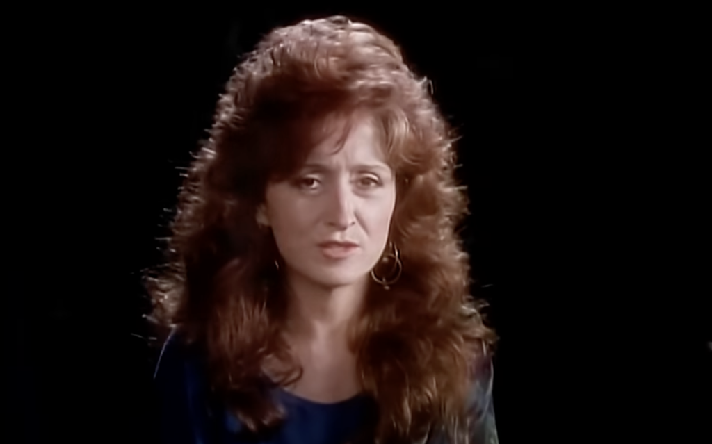 A woman with curly reddish-brown hair, wearing gold hoop earrings and a dark-colored top, is looking forward against a black background.