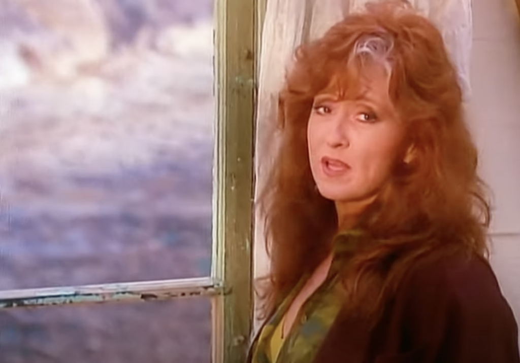 A person with long, wavy red hair is standing beside a partially open window, looking towards the camera. The background shows a blurred outdoor scene with soft natural lighting. The person is wearing a dark jacket over a patterned shirt.