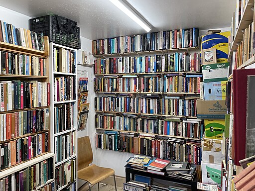 A small, cozy room filled with shelves of books lining the walls. There is a wooden chair in one corner and various boxes piled up on the right side. The bookshelves are organized with a wide variety of titles, creating a library-like atmosphere.