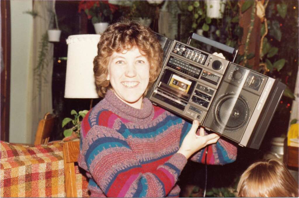 A woman with curly hair, wearing a colorful striped sweater, smiles as she holds a large vintage boombox on her shoulder. The background shows indoor plants and home decor, including a plaid-patterned fabric and hanging light fixture.