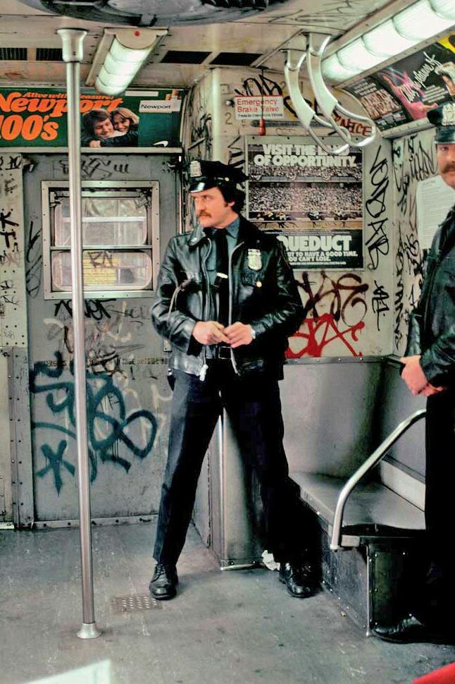 A police officer in a black leather jacket stands inside a heavily graffiti-covered subway car, holding onto a vertical pole. Another officer, partially visible, stands near the exit door. Subway ads are visible on the car’s walls.