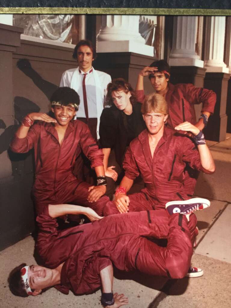 A group of six people, five men and one woman, pose in front of a classical building with columns. The men, wearing matching red jumpsuits and headbands, strike various dynamic poses, while the woman, in dark attire, stands in the middle smiling.