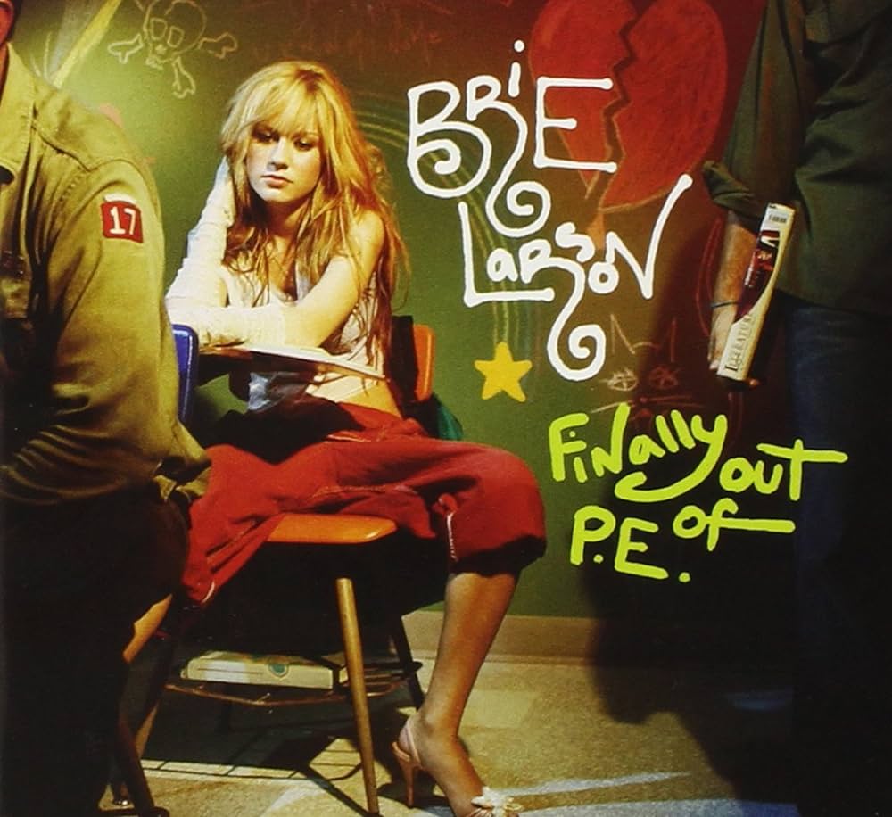 A young woman with long blonde hair, wearing a white sleeveless top and red pants, sits on a school desk with one leg resting on the seat. Behind her, colorful graffiti spells out "Brie Larson" and "Finally Out of P.E." on a chalkboard.