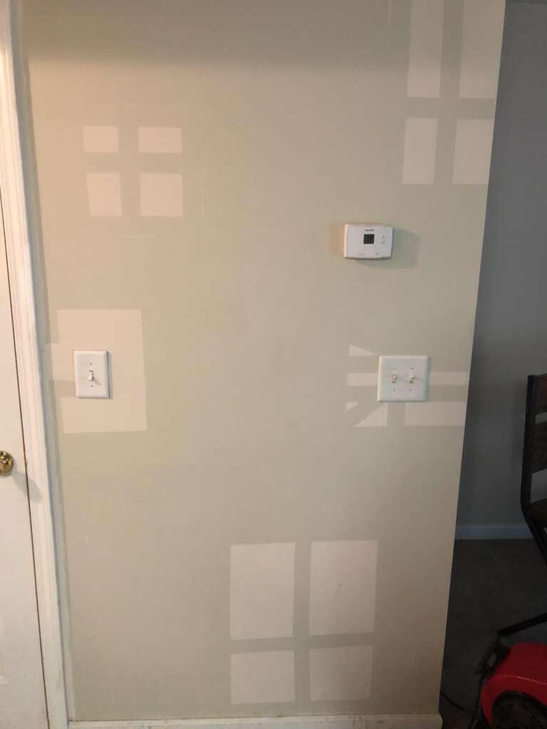 A beige wall with several patches of differently colored squares and rectangles indicating repair or repainting in progress. The wall features a light switch, a thermostat, and another switch or socket cover. A portion of a door frame and part of a chair are visible on the sides.