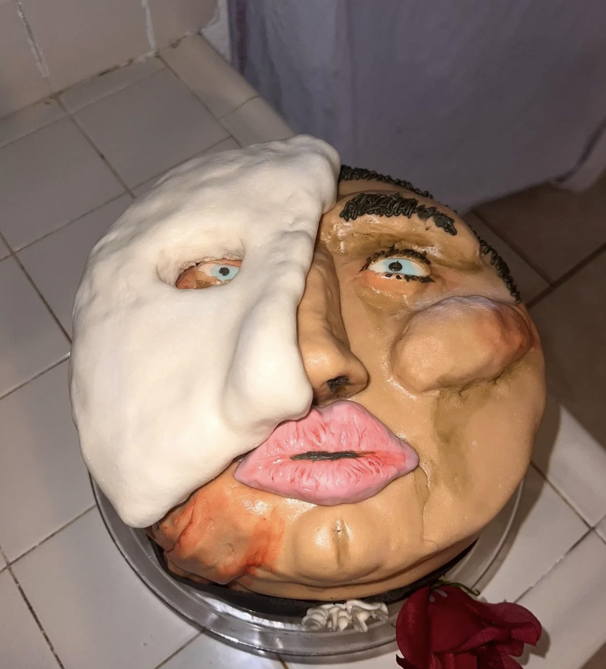 A cake decorated with a human face, half of which is covered with a creamy white mask. The face has detached, realistic features like eyes, lips, and a nose. The cake sits on a tiled surface, and a rose lies beside it.