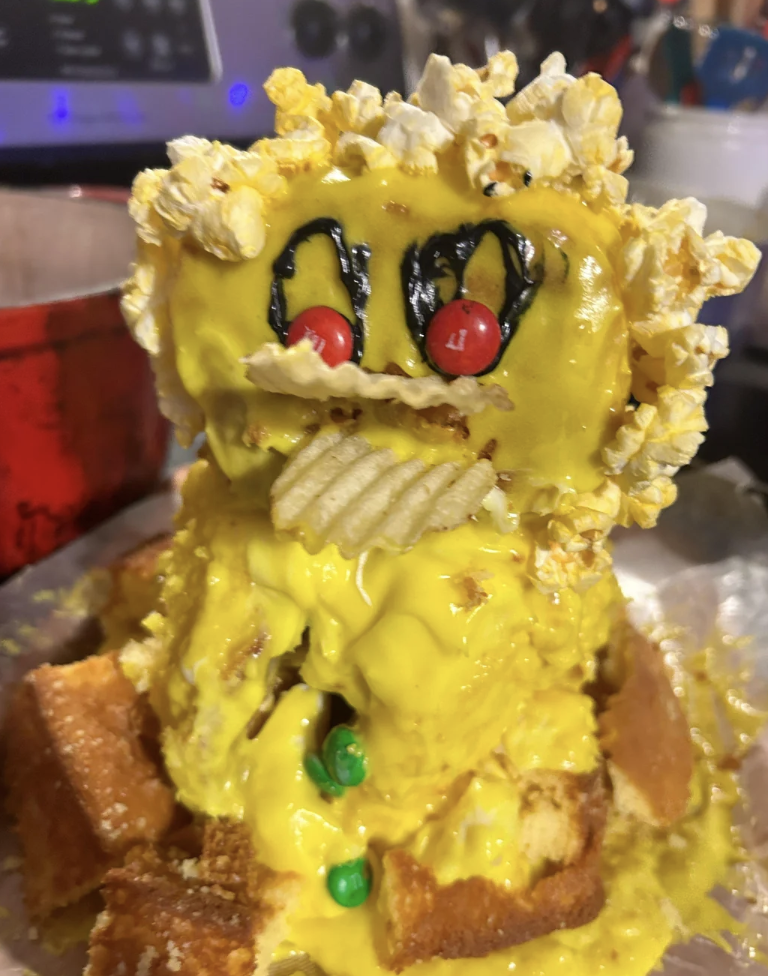 A quirky dessert creation featuring a sponge cake sculpture decorated to look like a character with yellow frosting, popcorn for hair, red candies for eyes, and a potato chip as a mouth. Colorful candies adorn the base of the cake.