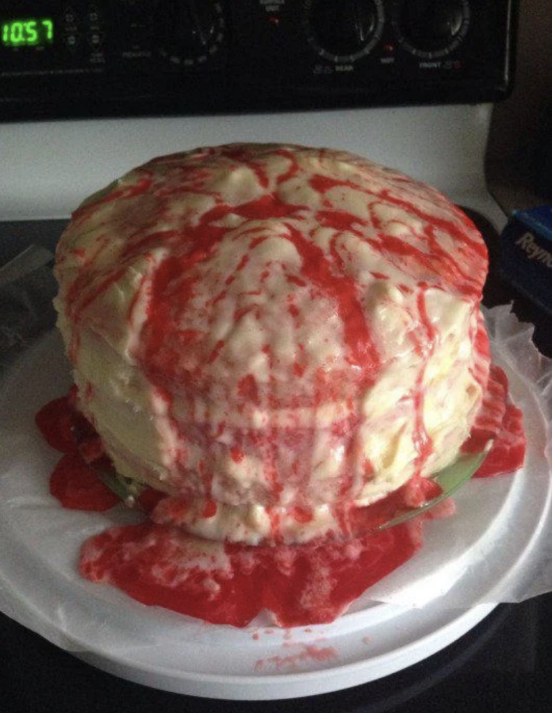 A round cake with white frosting and red-colored syrup drizzled over it, giving it a messy appearance. The syrup has pooled at the base, creating a striking contrast with the white frosting. The cake is placed on a white plate with a plastic wrap underneath.