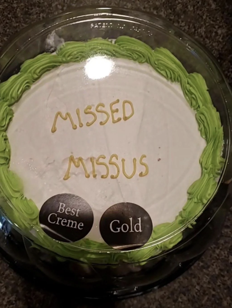 A round cake with white frosting and green decorative edging. The center has "Missed Missus" written in yellow icing. Two black stickers on the cake plastic lid read "Best Creme" and "Gold." The background is a dark, textured surface.