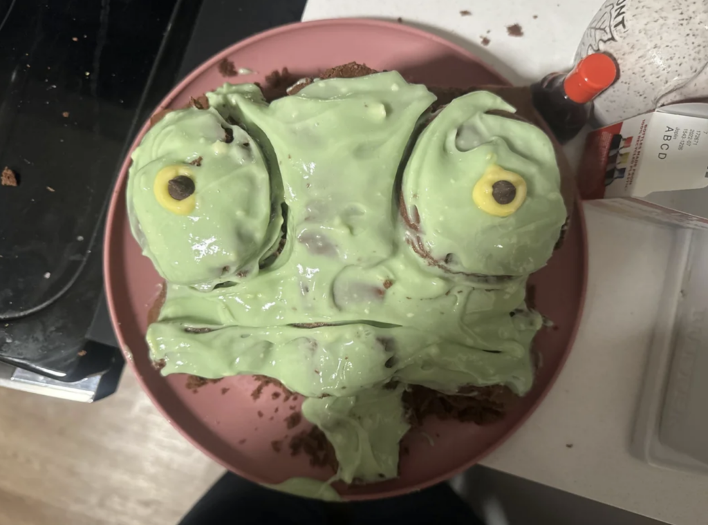 A cake designed to look like a frog's face on a pink plate. The cake has green frosting for the frog's skin and small yellow frosting with brown dots for the eyes. It sits on a kitchen counter with some baking supplies visible nearby.