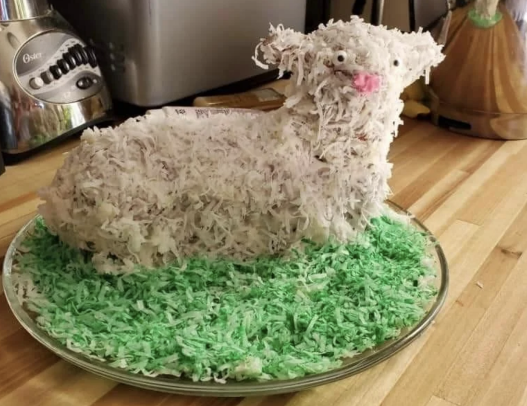 A cake shaped like a lamb covered in white shredded coconut sits on a glass plate. The plate is sprinkled with green shredded coconut to resemble grass. The lamb has a pink nose and black eyes, and there are kitchen appliances in the background.