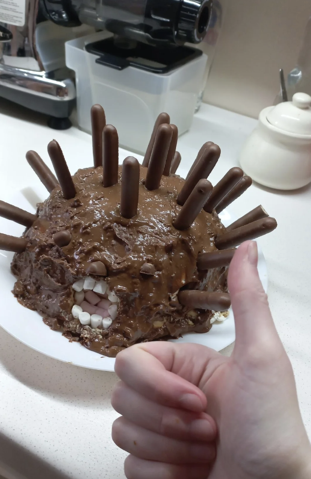 A round, chocolate cake decorated to look like a quirky monster with chocolate sticks as spikes, white candy teeth, and two chocolate pieces for eyes. A hand with a thumbs-up gesture is in the foreground, indicating approval of the cake.