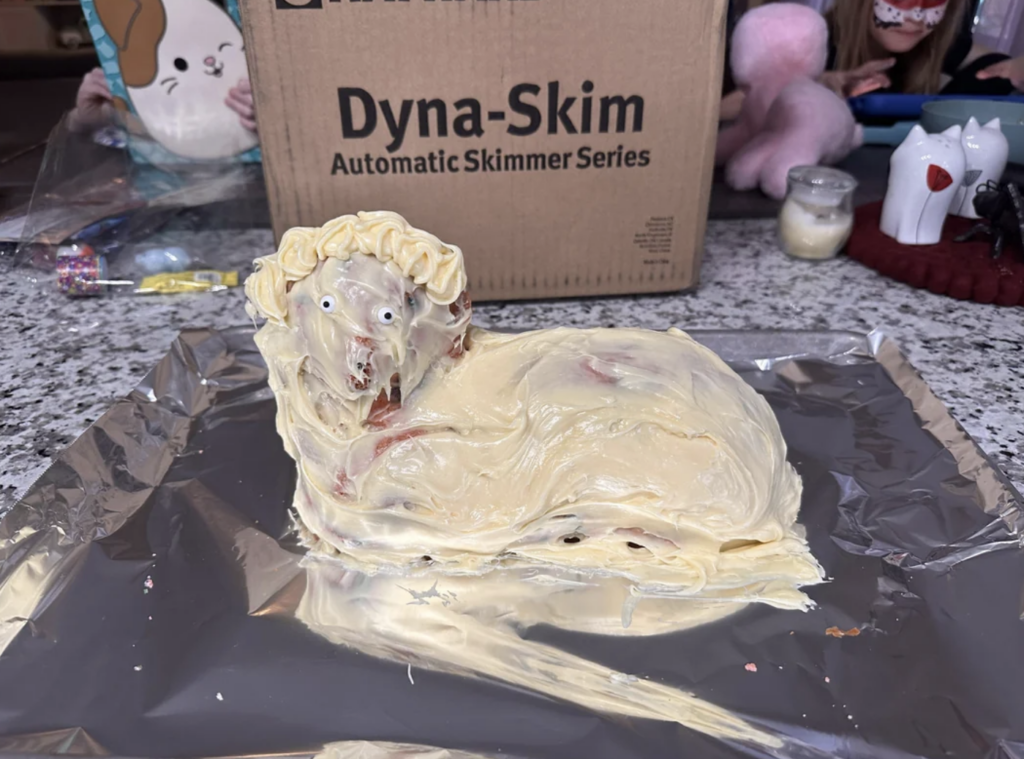 A homemade cake shaped like a lion rests on a sheet of aluminum foil. The lion cake is covered in cream-colored frosting, with distinct eyes and a mane decorated with swirls of icing. In the background, there is a cardboard box and various stuffed animals.