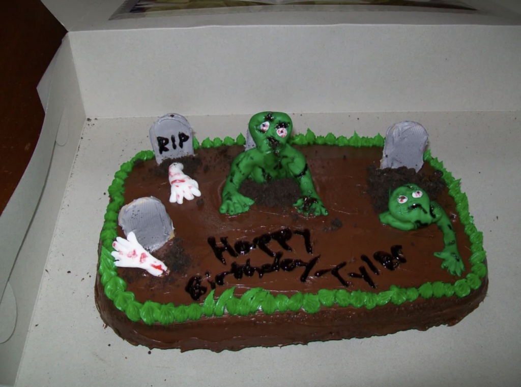 A birthday cake designed as a graveyard scene features green zombie figures emerging from the ground, alongside tombstones marked "RIP." The cake, decorated with green frosting grass, reads "Happy Birthday Tyler" in black icing on a brown chocolate surface.