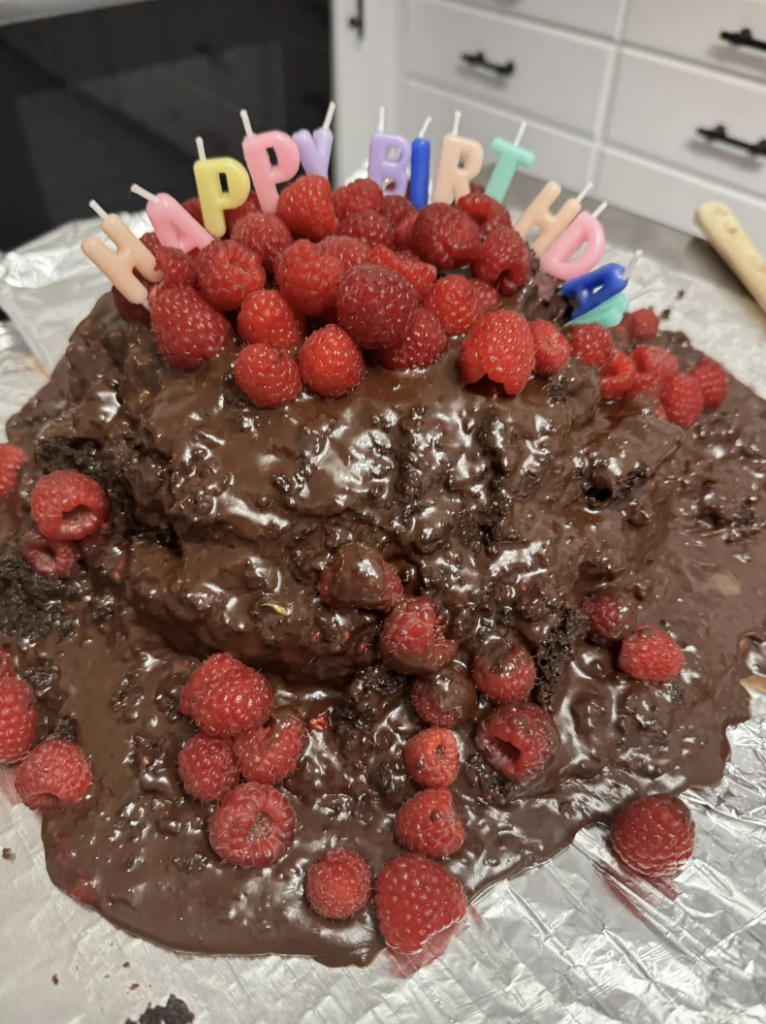 A homemade birthday cake covered in chocolate ganache and topped with fresh raspberries. Colorful candles spelling "HAPPY BIRTHDAY" are placed on top of the cake. The cake appears slightly messy with chocolate and raspberries spread around.