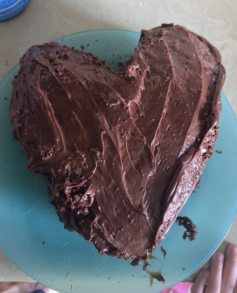 A homemade heart-shaped chocolate cake with textured chocolate frosting is placed on a teal plate. The surface of the cake is uneven, adding a rustic appearance. The edge of a pink object is visible at the bottom right corner.