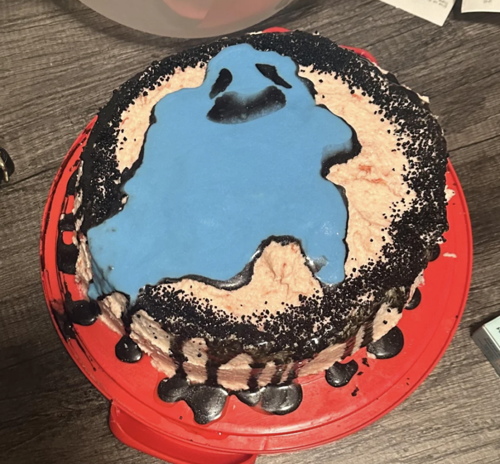 A homemade cake sitting on a red base, decorated with a blue ghost shape on top. The ghost has black eyes and mouth, and the cake's edges are sprinkled with black crumbs. Drips of black icing run down the sides. The wooden table background is slightly visible.