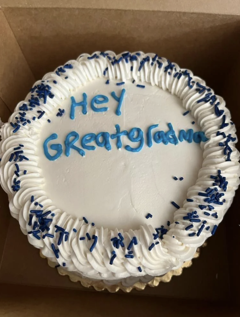 A round cake with white frosting, decorated with blue sprinkles around the edges. Written in blue icing on the top are the words "HEY GREATGRADMAS" with some letters appearing slightly smudged. The cake is in an open brown box.