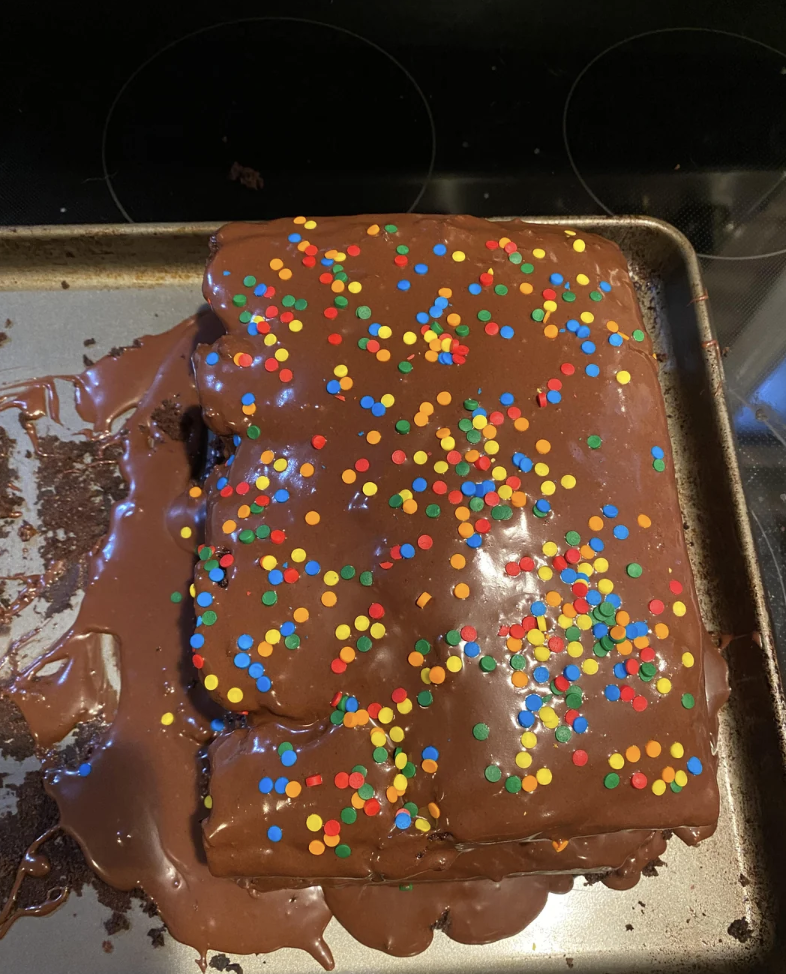 A homemade, rectangular chocolate cake with frosting spilling over the sides sits on a baking sheet. The top is decorated with colorful sprinkles, creating a festive appearance. The cake appears slightly uneven with some frosting melted and pooled at the base.