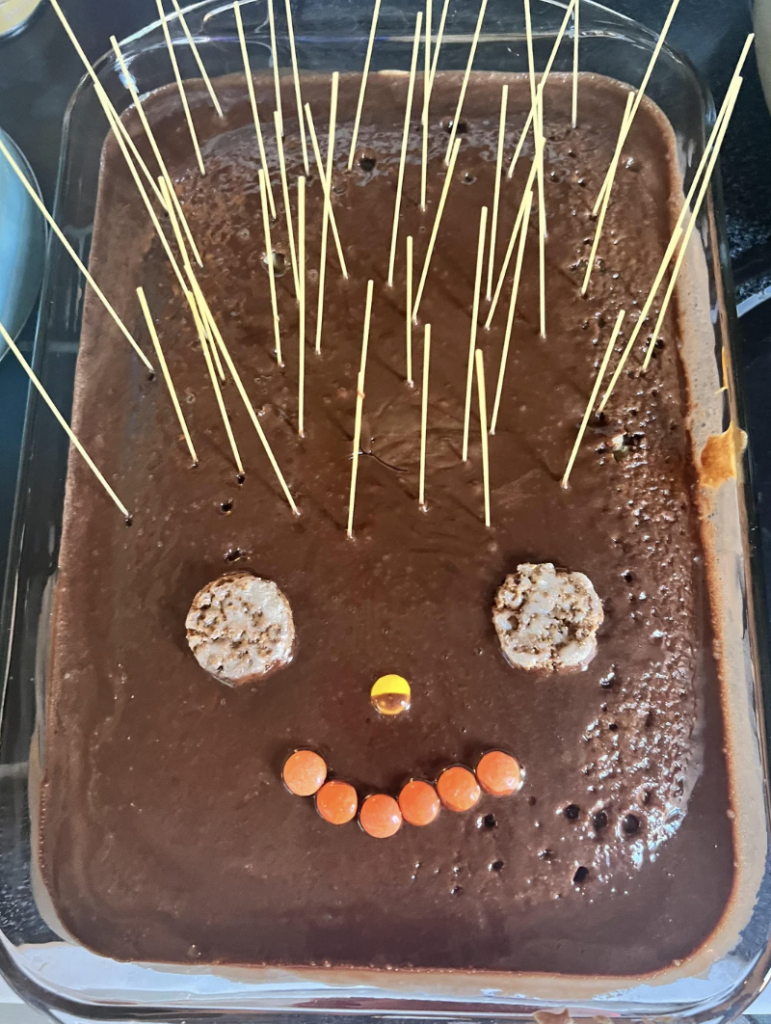 A rectangular chocolate cake is decorated to look like a face with two round cereal pieces for eyes, a yellow candy piece for a nose, and a smile formed from orange candy pieces. Uncooked spaghetti noodles are sticking out from the top of the cake like hair.