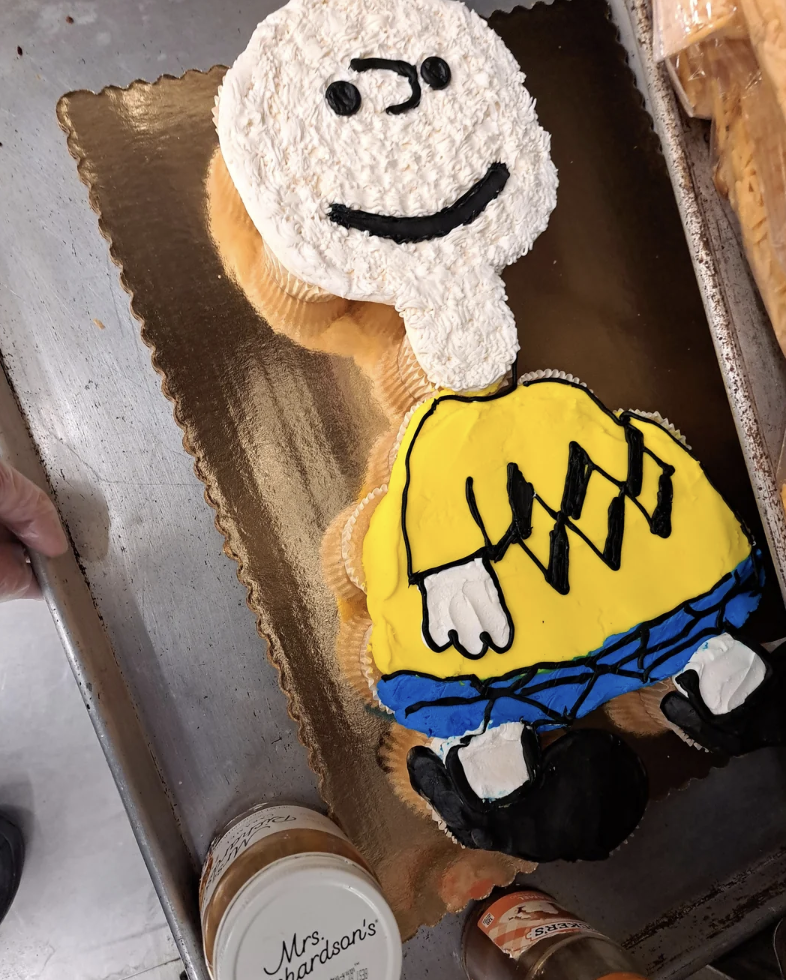 A large cupcake cake arranged to form a character with a smiling face and a yellow shirt with a black zigzag pattern. The cake is placed on a golden board, with jars of frosting nearby.