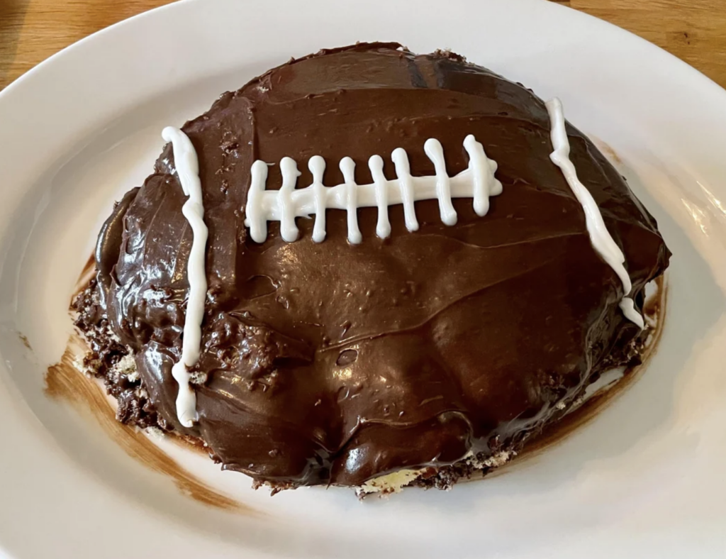 A chocolate cake shaped like a football is displayed on a white oval plate. The cake is frosted with chocolate icing and decorated with white icing to resemble the laces and stitches of a football. The bottom edges of the cake show some roughness.