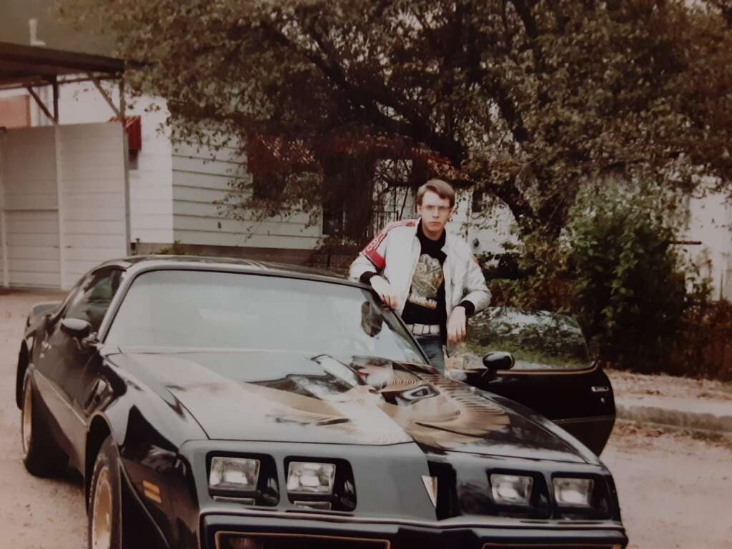 A man stands beside a black Pontiac Firebird Trans Am with its door open. He is wearing a light-colored jacket over a graphic t-shirt and jeans. The car has a large decal of a firebird on its hood. Trees and a building can be seen in the background.