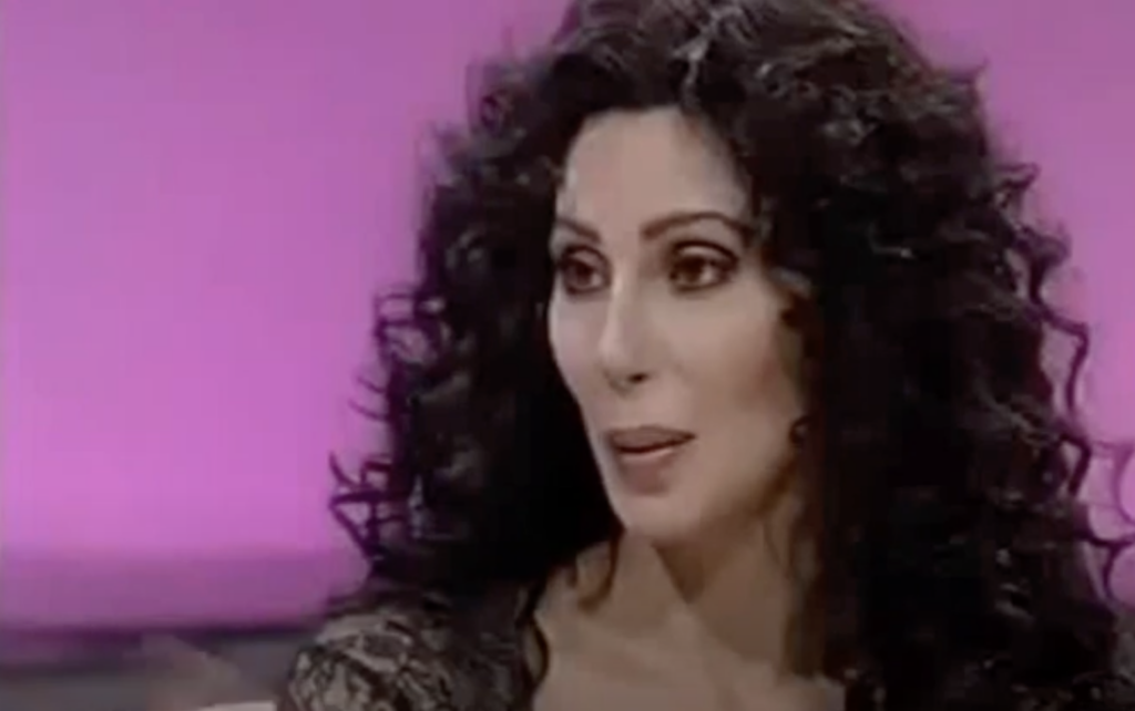 Person with long, curly black hair and light makeup, wearing a black top, appears to be speaking or reacting during what looks like an interview or talk show. The background is a pink, softly-lit setting.