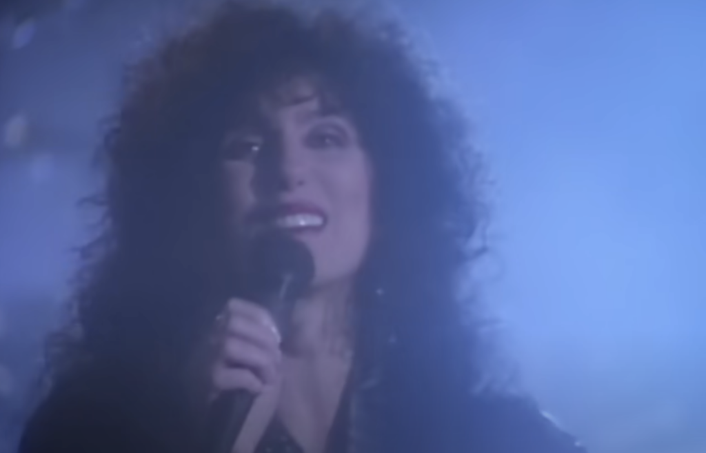 A person with curly hair is singing into a microphone against a hazy, blue-lit background. The person is smiling and looking slightly upward as they perform.