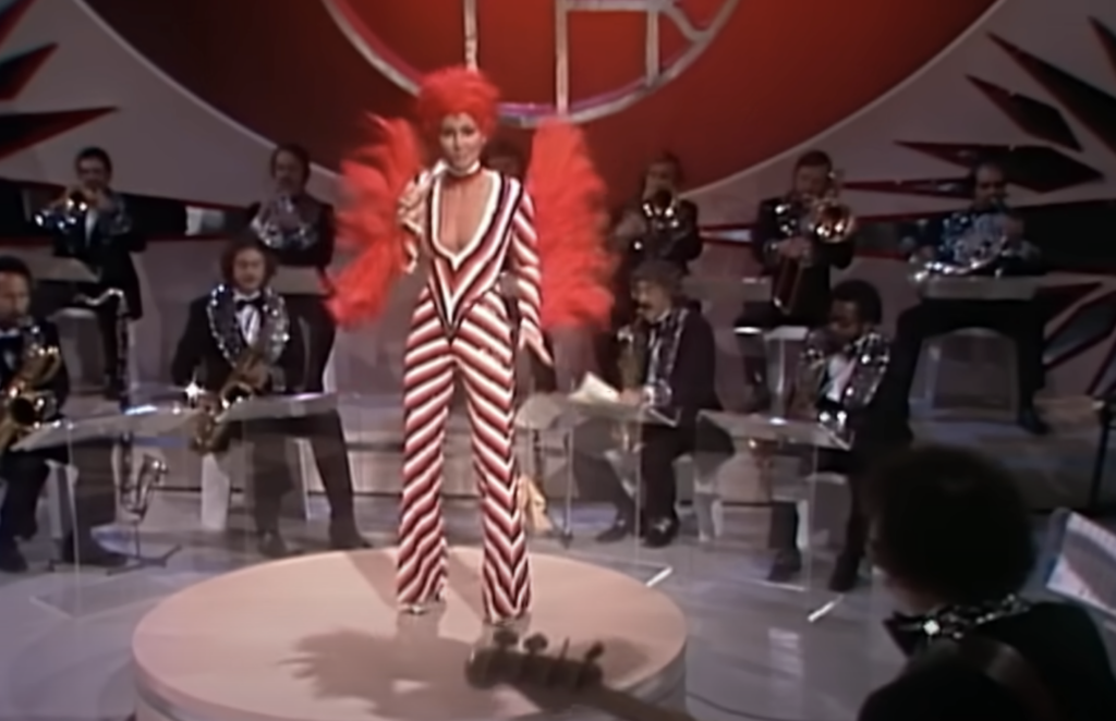 A person stands on a circular stage wearing a red and white striped jumpsuit with a plunging neckline and vibrant red headpiece. They are surrounded by seated musicians playing various instruments, performing under dramatic lighting and a stylized backdrop.