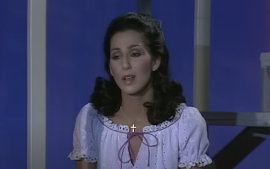 A woman with long dark hair wearing a light purple blouse with a bow and a cross necklace is seated against a blue background, and she appears to be singing or speaking.