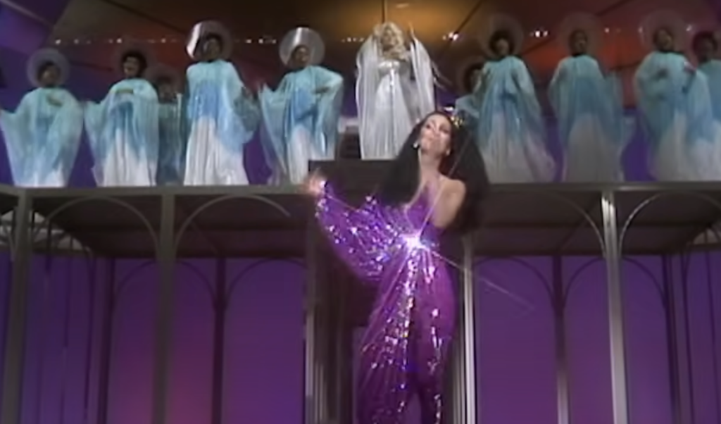 A singer wearing a sparkly purple dress performs on a stage with arms outstretched, while a group of background singers dressed in blue and white robes stand elevated behind her. The background features vibrant, colorful lighting.