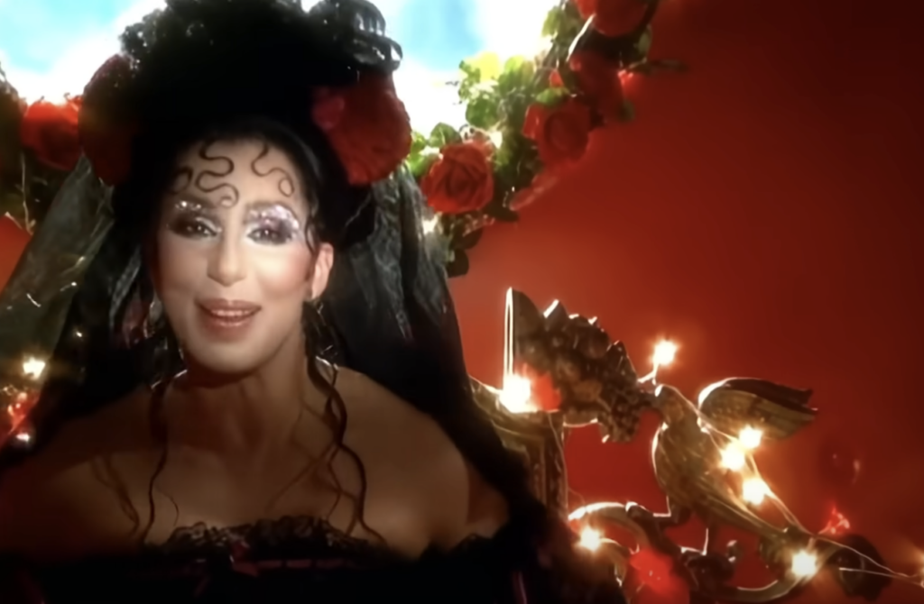 A person with elaborate makeup, wearing a black outfit and a headpiece decorated with red flowers and a black veil, smiles in front of a red background adorned with roses and bright lights.