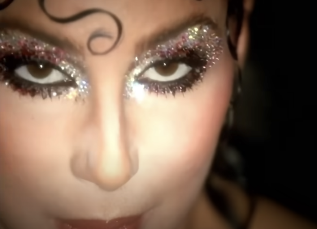 Close-up of a person’s face with glittery, dramatic makeup. Their eyelids are covered in sparkling glitter, and a curl of hair is visible on their forehead. The expression is intense, with eyes partly closed and lips softly pursed.
