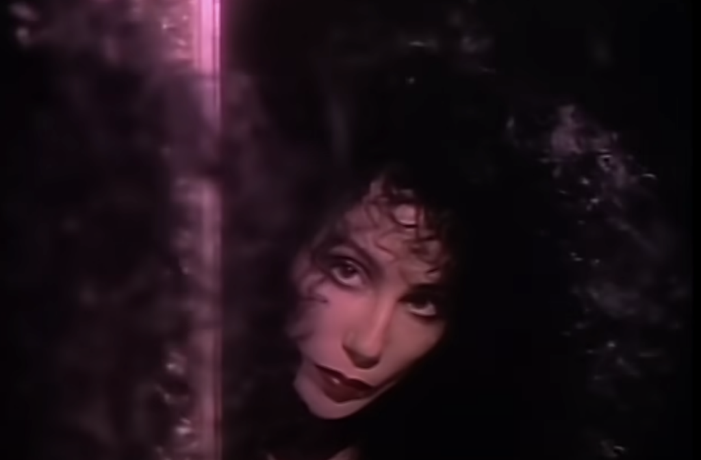 A person with voluminous curly dark hair and dramatic makeup is pictured in low lighting, gazing towards the camera through a dimly lit reflective surface. Their expression is intense and the background is largely dark, adding an air of mystery.