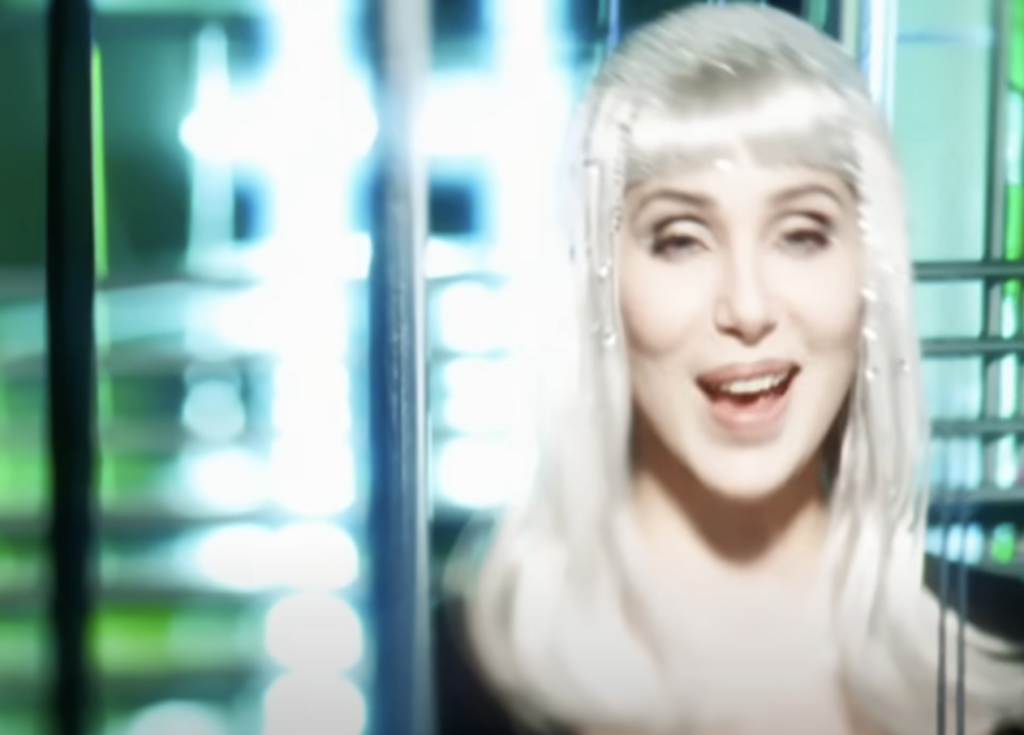 A person with long silver hair and wearing a dark top is smiling in a futuristic setting with bright, blurred lights in the background.