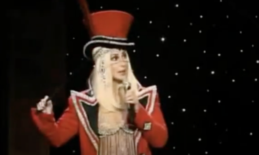 A person wearing a red ringmaster-style costume with an elaborate top hat and decorative embellishments stands against a dark, starry background. They hold a baton in one hand and appear to be mid-speech or performance.