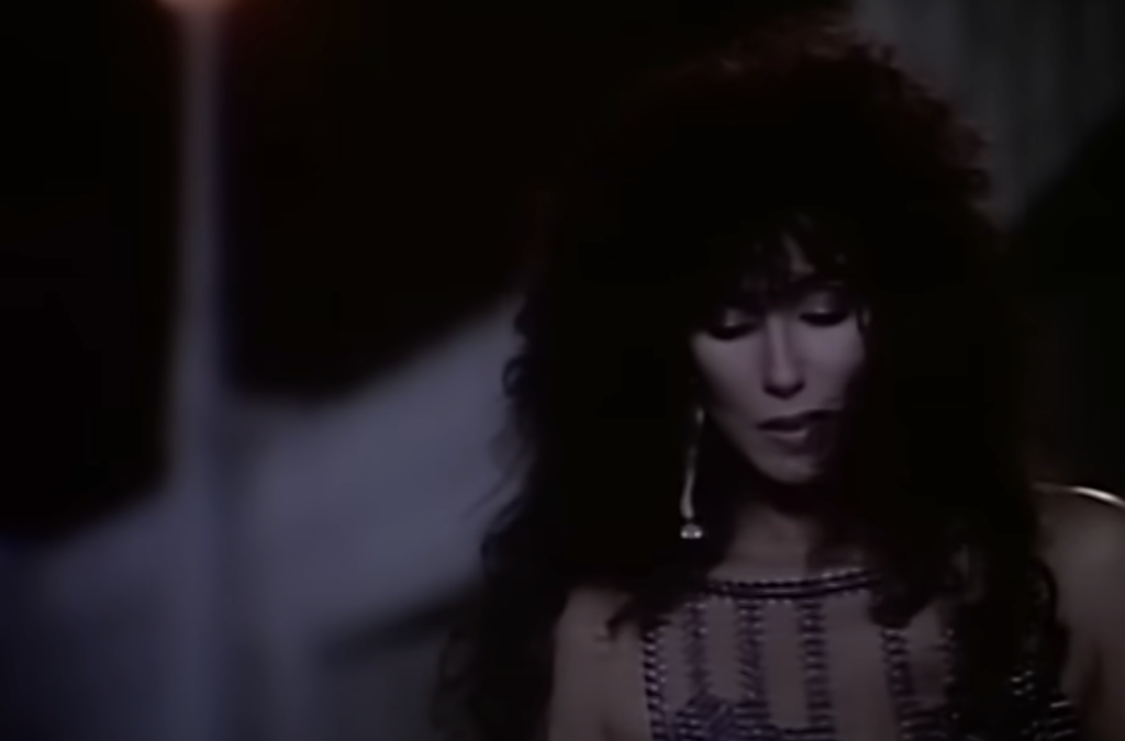 A person with long, curly hair is shown in a nighttime setting. They are dressed in a beaded outfit with an intricate neckline and are looking downward. The background is dimly lit with a soft orange light, creating a moody atmosphere.