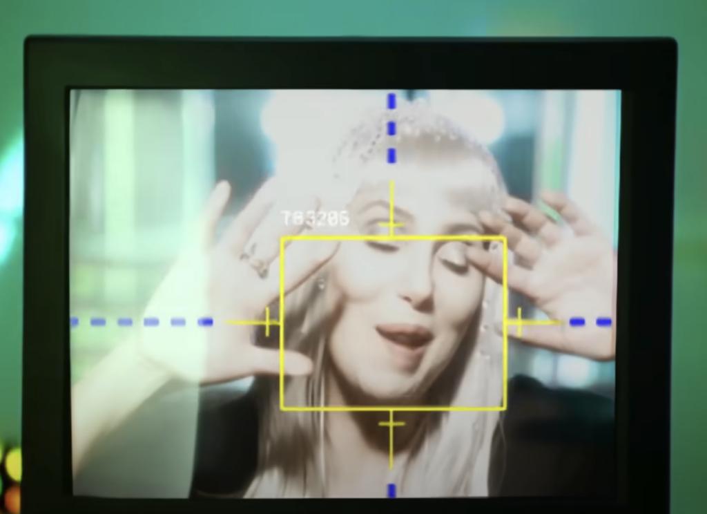 A person with long blonde hair is shown on a screen, framed with yellow and blue guidelines. They have their eyes closed, mouth slightly open, and hands raised near their face, creating a playful expression. The background appears blurred.