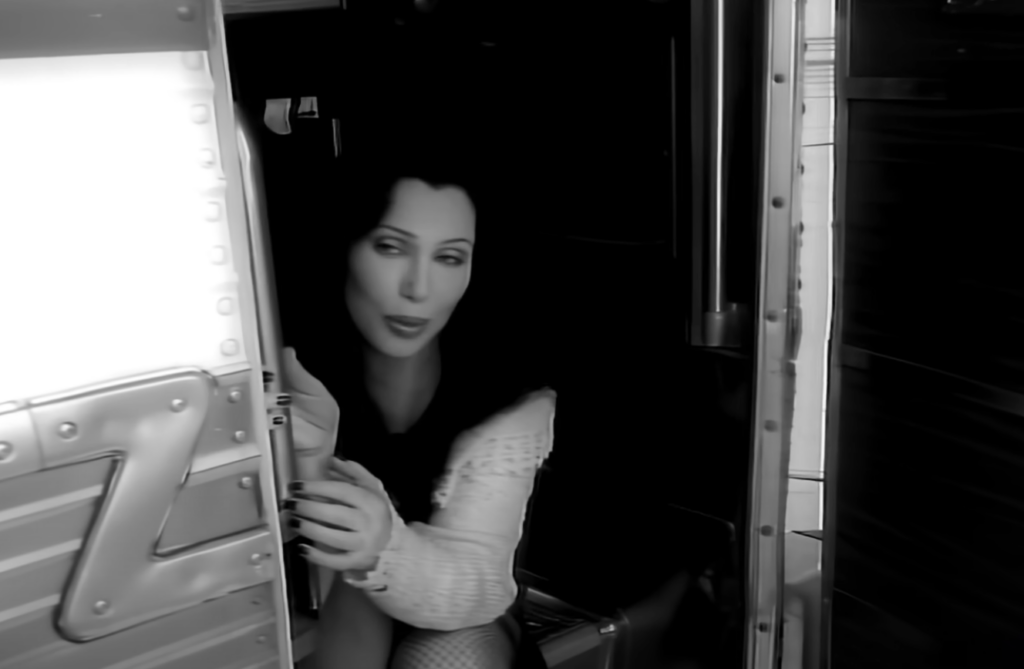 A person with long, dark hair and wearing a light-colored top is partially visible, emerging from the doorway of a truck. They have a focused expression and are holding onto the truck's door with one hand, looking ahead through the open door.
