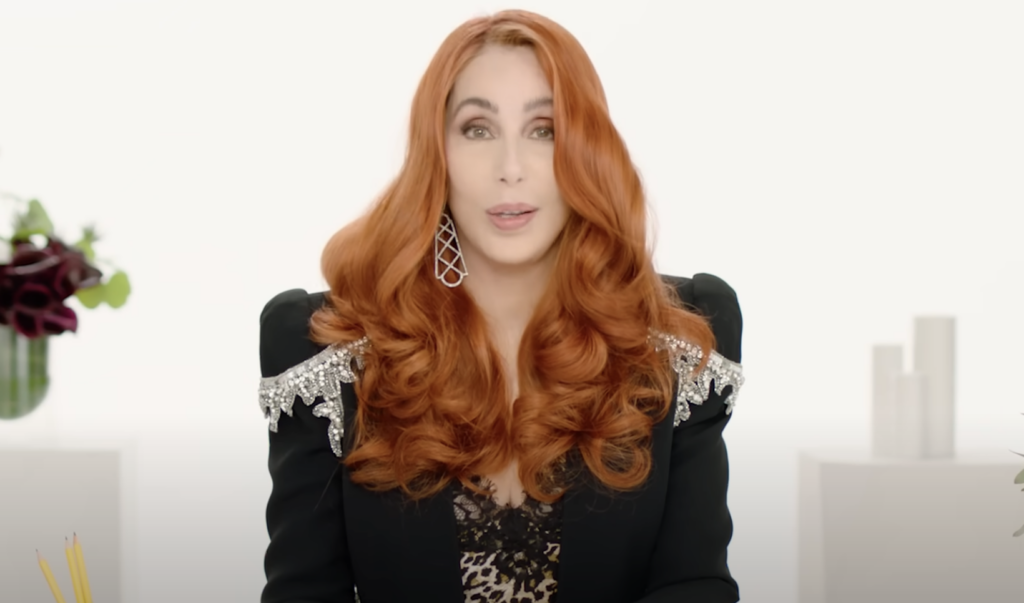 A person with long, curly red hair is sitting against a white background. They are wearing a black outfit with sequin details on the shoulders and a patterned top underneath. They have large, dangling earrings and are looking towards the camera.